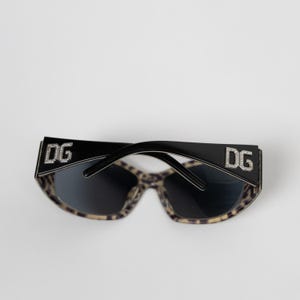 May include: Black sunglasses featuring a leopard print pattern on the inner frame and dark lenses. The wide temples are adorned with the letters DG in a silver-toned finish, embellished with small clear crystals on each side of the frame.