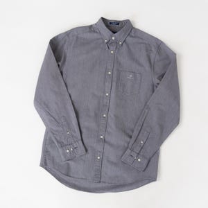 May include: A long-sleeve button-down shirt in a solid dark gray color. The shirt features a pointed collar, a single chest pocket on the left side with a small embroidered logo, and buttoned cuffs. The fabric has a subtle, fine-textured weave.
