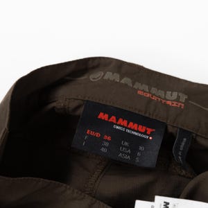 Puede incluir: Primer plano de pantalones de senderismo marrones con la marca "MAMMUT" visible. La etiqueta muestra la talla EU 36, UK 10 y USA 6. Los pantalones est&aacute;n hechos en China.