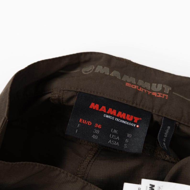 Puede incluir: Primer plano de pantalones de senderismo marrones con la marca "MAMMUT" visible. La etiqueta muestra la talla EU 36, UK 10 y USA 6. Los pantalones est&aacute;n hechos en China.