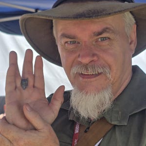 May include: A man with a white beard and mustache is wearing a brown felt hat with a black band. He is wearing a green shirt and has a badge on his chest. He is holding his hand out with a small, dark object on his finger.