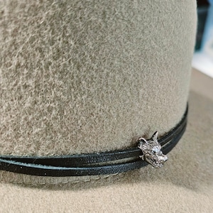 May include: A sage green fedora hat with a black leather band. The hat features a silver-colored wolf head charm on the band. The hat is made of a soft, felt-like material.