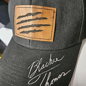 Könnte beinhalten: Schwarze Baseballkappe mit einem braunen Lederpatch mit einem Krallenmuster. Die Kappe ist mit "Blackie Thomas" in Weiß signiert.