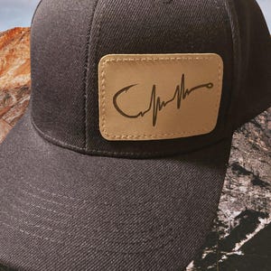 May include: A black baseball cap with a brown leather patch featuring a fishing hook and heartbeat design. The patch has the text "Catch" on it.