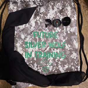 May include: A black and white camouflage drawstring bag with the text "FUTURE SILVER WOLF IN TRAINING" in green letters. The bag has a black fabric lining and a compass attached to the front.