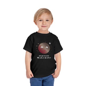 Puede incluir: Una camiseta negra con una ilustración de dibujos animados del planeta enano Makemake. El texto "Planeta enano Makemake" está impreso debajo de la ilustración.