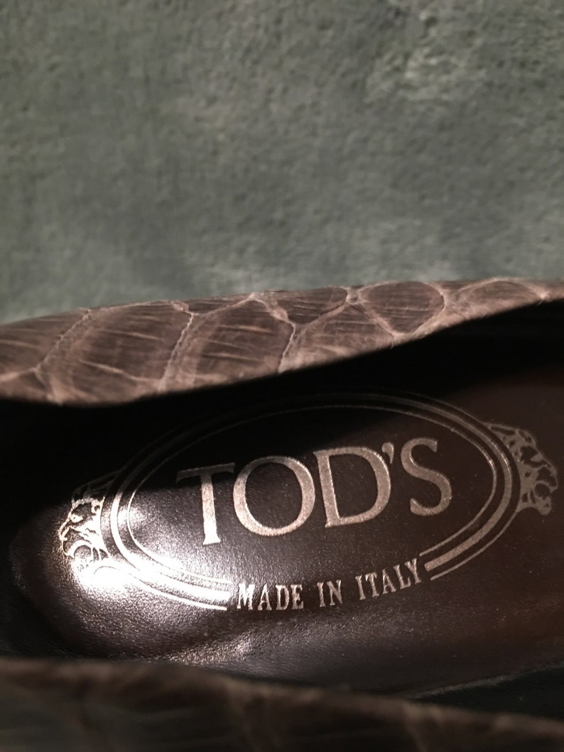Op de afbeelding: Close-up van de binnenkant van een donkerbruine leren schoen. De binnenzool toont de merknaam "TOD'S" in een zilveren ovaal, met "MADE IN ITALY" eronder. De buitenkant van de schoen heeft een gestructureerd, schubachtig patroon.