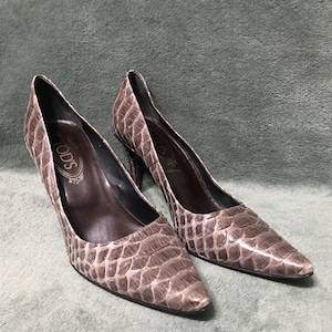 Può includere: Un paio di décolleté Tod's con tacco alto. Le scarpe sono di colore talpa neutro con una texture simile a quella di un rettile. Hanno punte a punta e tacchi sottili. L'interno è di colore marrone scuro.