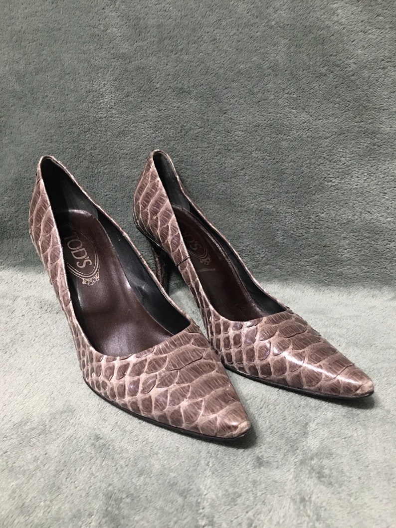 Op de afbeelding: Een paar Tod's pumps met hoge hakken. De schoenen hebben een neutrale taupe kleur met een reptielachtige textuur. Ze hebben spitse tenen en slanke hakken. De binnenkant is donkerbruin.