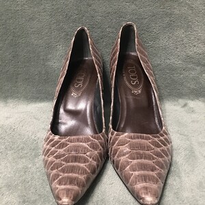 Op de afbeelding: Een paar bruine pumps met spitse neus en een slangenleerpatroon. De hakken zijn middelhoog en de binnenkant is donkerder bruin. De merknaam "TOD'S" is zichtbaar in de schoen.