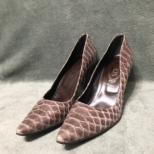 Op de afbeelding: Een paar grijze pumps met spitse neus en een reptielachtige textuur. De hakken zijn middelhoog en de binnenkant is donkerbruin. De merknaam "TOD'S" is zichtbaar in de schoen. De schoenen staan tegen een gedempte groene achtergrond.