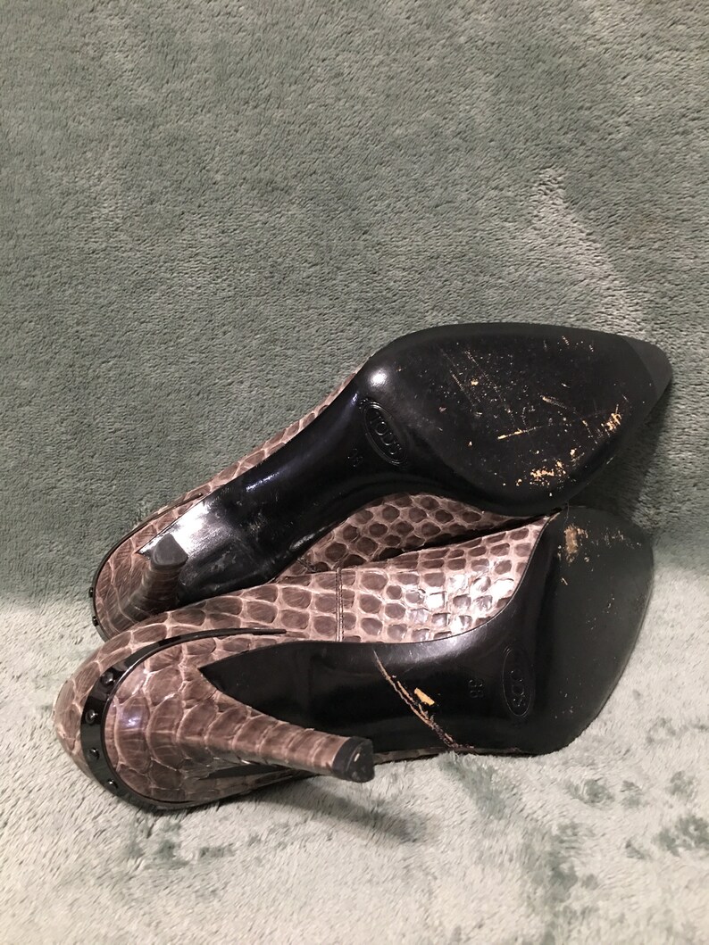 Op de afbeelding: Een paar pumps met een slangenprint in bruin- en grijstinten. De hakken zijn zwart en de zolen vertonen slijtage. De schoenen hebben een spitse neus en een decoratief metalen detail op de hiel.