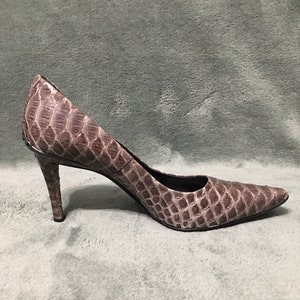 Op de afbeelding: Een enkele, puntige high heel schoen. De schoen heeft een neutrale kleur met een textuur, reptielachtig patroon. De hak is dun en de schoen staat tegen een effen achtergrond.