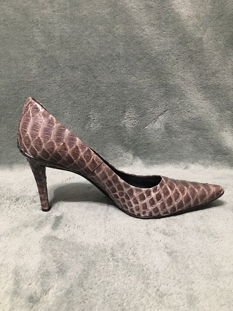 Op de afbeelding: Een enkele, puntige high heel schoen. De schoen heeft een neutrale kleur met een textuur, reptielachtig patroon. De hak is dun en de schoen staat tegen een effen achtergrond.