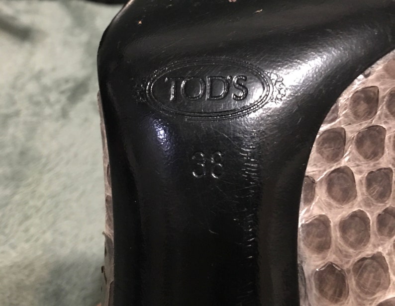 Op de afbeelding: Close-up van de zool van een pumps. De zwarte zool is voorzien van het merk "TOD'S" en maat "38". De schoen heeft een slangenprint in grijstinten en beige. Maat 38.