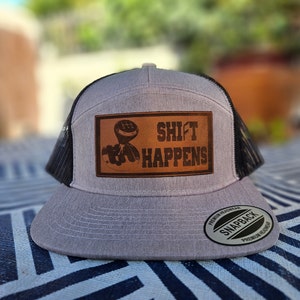 Peut inclure: Un chapeau de camionneur gris et noir avec un patch en cuir marron qui dit "Shift Happens" et un graphique d'un levier de vitesses. Le chapeau a un dos en maille noire et une fermeture à pression noire.