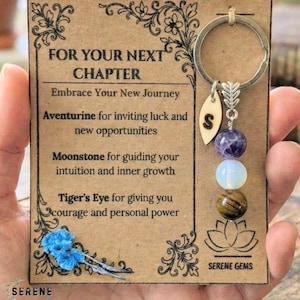 May include: A keychain with a silver ring, a wooden leaf charm with the letter "S", and three gemstone beads: purple, white, and brown. The keychain hangs from a card that reads "FOR YOUR NEXT CHAPTER" and lists the properties of each gemstone.