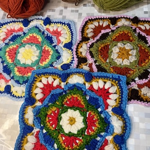 May include: Close-up of three colorful, square crochet pieces with intricate floral patterns. The pieces feature a variety of colors, including blue, red, yellow, green, and white. Several balls of yarn in shades of brown, orange, and green are visible in the background.