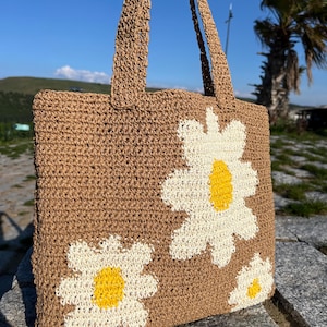May include: A brown woven tote bag with white and yellow daisy flower appliqués. The bag has two handles and is made of a natural fiber material. The daisies are in various sizes and are placed on the front of the bag.