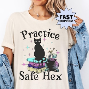 Puede incluir: Camiseta color crema con el texto "Practice Safe Hex" y un gráfico de un gato negro sobre libros, un caldero y estrellas. La camiseta es una camiseta Bella+Canvas.