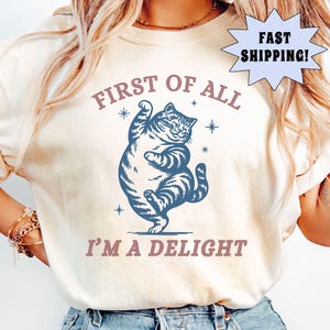 May include: Ivory-colored t-shirt featuring a blue cartoon cat with the text "FIRST OF ALL I'M A DELIGHT". The cat is standing on its hind legs with one paw raised. The shirt has a relaxed fit and is made of a soft material.