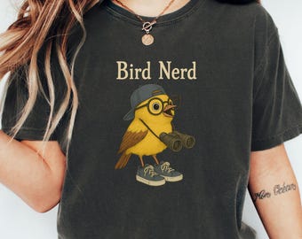 May include: Dark gray t-shirt featuring a cartoon bird wearing a cap, glasses, and sneakers, holding binoculars. The text "Bird Nerd" is printed above the bird illustration. The shirt is a comfortable, casual style.