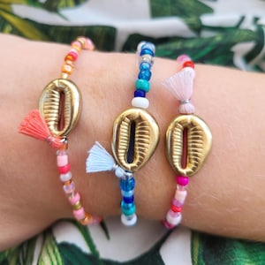 Op de afbeelding: Drie kleurrijke kralenarmbanden met gouden kauri schelp bedels. De armbanden zijn gemaakt van roze, blauwe en rode kralen.