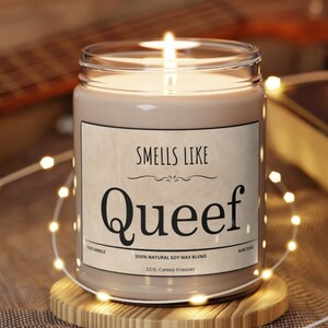 May include: A brown soy wax candle with a white label that reads "SMELLS LIKE Queef" and "100% NATURAL SOY WAX BLEND" and "LOL CANDLE GALLERY". The candle is lit and surrounded by a string of white lights.