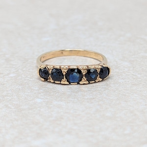 May include: A gold ring featuring five round, dark blue gemstones set in a row. The band is a simple, polished gold, and the gemstones are evenly spaced, creating a classic and elegant design. The ring is likely a piece of jewelry.