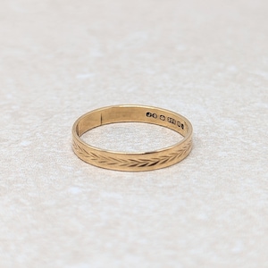 May include: A gold band ring with a detailed leaf pattern etched around the outside. The ring has a smooth, polished finish and is likely made of gold. The ring is a classic piece of jewellery.