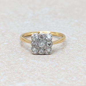 May include: A gold ring with a square cluster of small, clear gemstones. The ring's band is a simple, thin gold. The gemstones are set closely together, creating a sparkling, square shape. The ring is set against a neutral background.