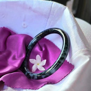 May include: A dark green jade bangle with white streaks, resting on a purple fabric with a white flower.