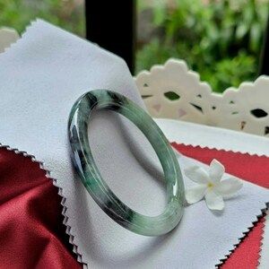 May include: A jade bangle bracelet with a mix of green and black hues. The smooth, polished surface reflects light, highlighting the natural variations in color. The bracelet is displayed on a white cloth with a red border and a white flower.