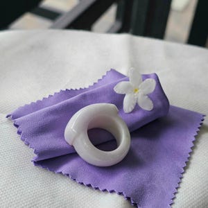 May include: A light-colored jade ring with a smooth, rounded design rests on a folded lavender cloth. A white flower with yellow center accents the scene. The ring's polished surface reflects light, highlighting its elegant form.