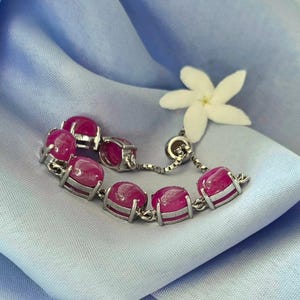 May include: A silver bracelet featuring oval-shaped, vibrant pink gemstones. The gemstones are set in a silver frame and linked together with a delicate chain. A white flower rests nearby on a light blue fabric.