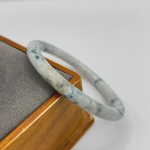 Puede incluir: Un brazalete de jade con aspecto veteado. El brazalete presenta una mezcla de tonos blancos, verdes y grises. La superficie lisa y pulida del brazalete se muestra contra una caja de madera.