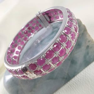 Op de afbeelding: Een zilveren armband met een rasterpatroon van ronde, roze edelstenen. De armband is scharnierend en lijkt van zilver te zijn. De edelstenen hebben een levendige roze kleur en de armband ligt op een lichtblauwe ondergrond.