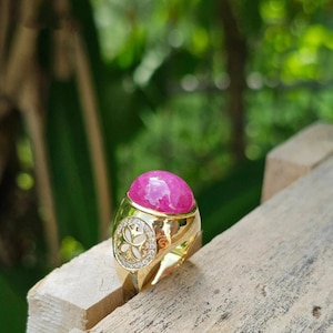 Op de afbeelding: Een gouden ring met een grote ovale roze edelsteen en een decoratief ontwerp van kleine diamanten rond de zetting.