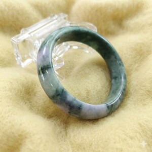 Op de afbeelding: Een jade armband met tinten groen en lavendel. Het gepolijste oppervlak reflecteert licht en benadrukt de natuurlijke kleurvariaties. De armband wordt gepresenteerd op een heldere, schuine standaard tegen een zachte, gele achtergrond.
