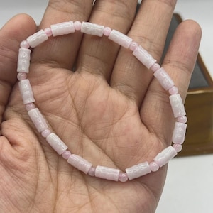 Op de afbeelding: Een delicate armband met afwisselend lichtroze en witte cilindrische kralen. De armband is geregen op een dun, elastisch koord, wat zorgt voor een flexibele en comfortabele pasvorm. De kralen hebben een gladde, gepolijste afwerking.