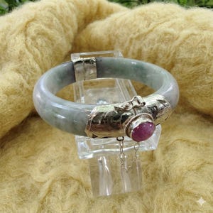 May include: A jade bangle bracelet with silver accents and a red gemstone. The bracelet is a light green color and has a clasp closure. The gemstone is round and set in a silver bezel.