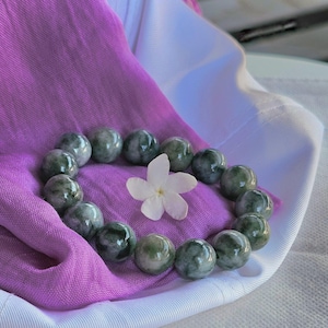 May include: A jade green beaded bracelet with a white flower on a purple and white fabric background.