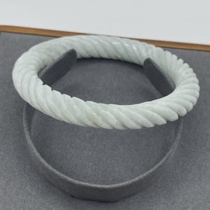 Op de afbeelding: Een lichtgroene jade armband met een gedraaid touwontwerp. De armband wordt getoond op een grijze standaard, die de ingewikkelde gebeeldhouwde textuur en bleke kleur benadrukt. De armband is een sieraad.