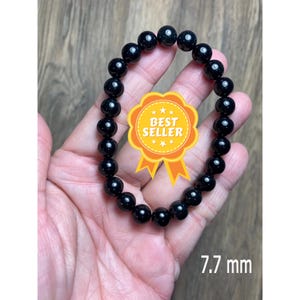 Könnte beinhalten: Schwarzes Perlenarmband mit einer Perlengröße von 7,7 mm. Das Armband besteht aus runden, schwarzen Perlen und wird an einem Handgelenk gezeigt.