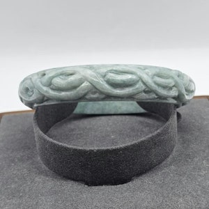 Puede incluir: Un brazalete de jade verde con un diseño tallado de vides entrelazadas. El brazalete es liso y pulido.