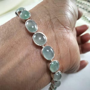 Op de afbeelding: Een zilveren armband met ovale, lichtgroene edelstenen. De stenen zijn gezet in individuele zilveren bezels en met elkaar verbonden. De armband wordt getoond op een pols, waarbij het ontwerp en de kleur worden benadrukt.