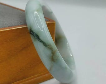 Könnte beinhalten: Ein Jade-Armreif in den Farbtönen Hellgrün und Weiß, mit Streifen aus dunklerem Grün und Braun. Die polierte Oberfläche reflektiert das Licht und hebt die natürlichen Variationen des Steins hervor. Dieses elegante Schmuckstück ist ein klassisches Accessoire.