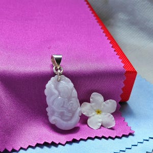 May include: A lavender-colored jade pendant with a silver bail, featuring a carved design. A white flower with a yellow center rests beside the pendant on a purple cloth. The background includes a red and blue fabric.