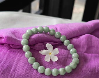 May include: A light green jade bracelet with round beads on a purple fabric background with a white flower.