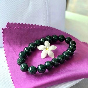Puede incluir: Un brazalete de jade verde oscuro con cuentas redondas sobre un fondo de tela morado. Una flor blanca descansa sobre el brazalete.